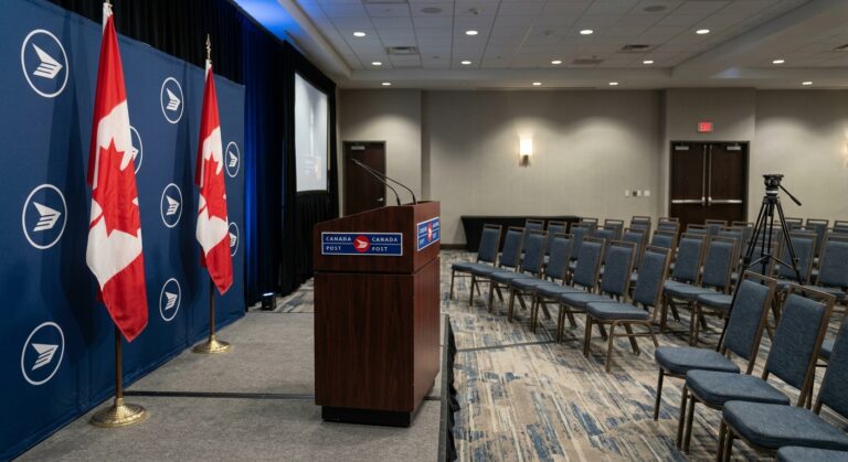 A professional photograph of an empty stage with two Canadian flags and a Canada Post branded backdrop, ready for a presentation about Canada Post Ecommerce Solutions.